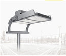 A large industrial outdoor stadium light fixture mounted on a pole, shown against a faded background of a sports court and fencing.